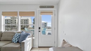 L'intérieur d'une maison de plage, avec une porte blanche, une vue sur la plage et les bâtiments, un canapé beige et un coussin bleu à motifs.