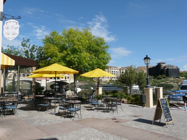 Un café al aire libre con sombrillas amarillas, mesas y sillas, con árboles y edificios de fondo.