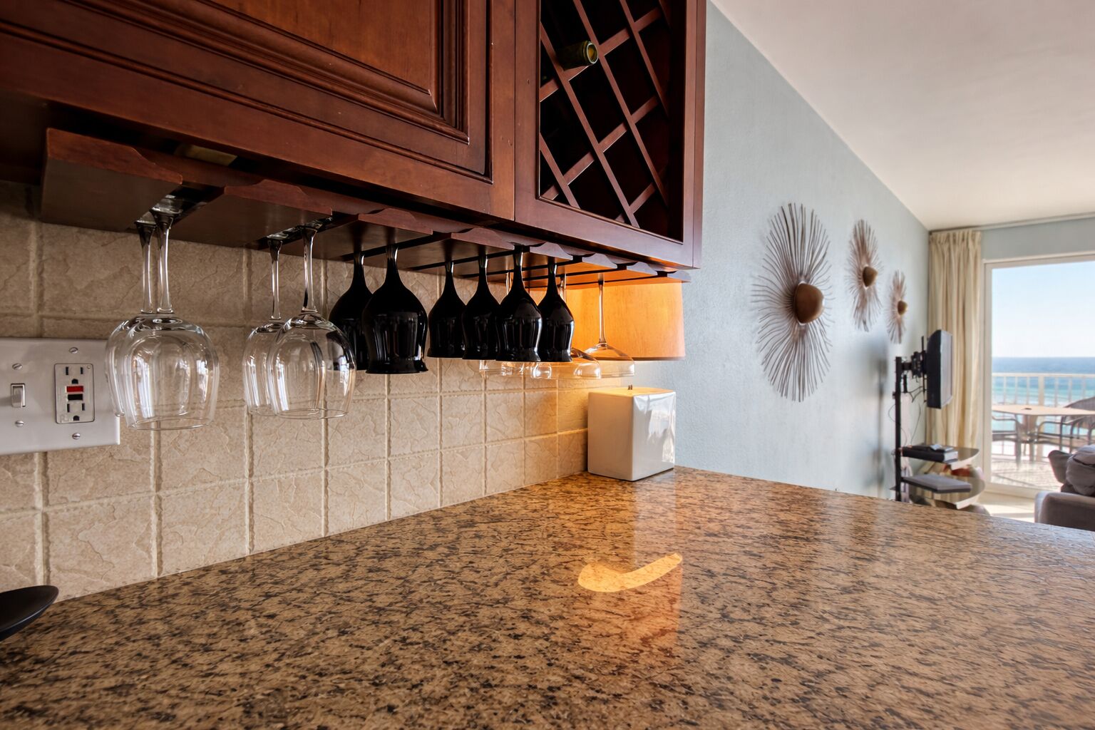 A kitchen with granite countertops, wine glasses hanging above, a wine rack, and a view of the ocean through a large window.