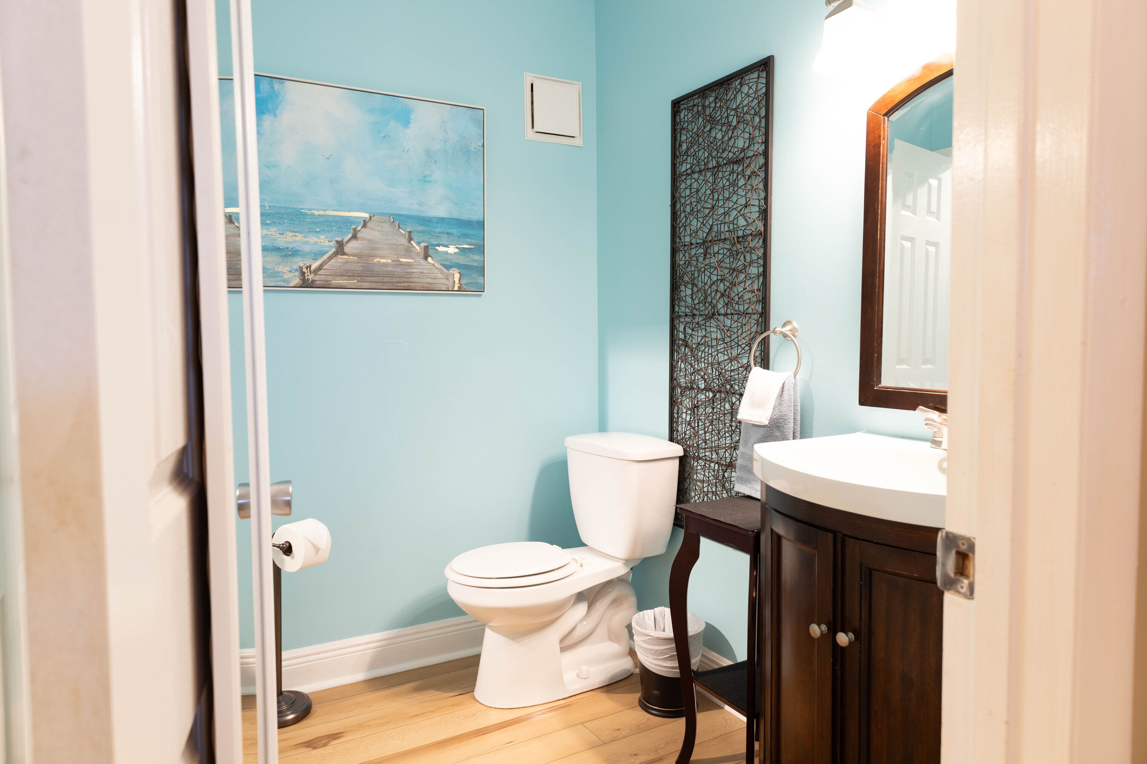 A bathroom with a white toilet, a wooden floor, a sink with a mirror, and a painting of a pier on the wall.