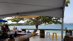 Un bar junto a la playa con vistas al mar, taburetes de madera y una mesa bajo una pérgola.