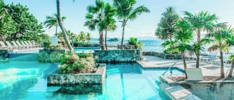 A swimming pool with clear water, surrounded by palm trees and lounge chairs, with a view of the ocean.
