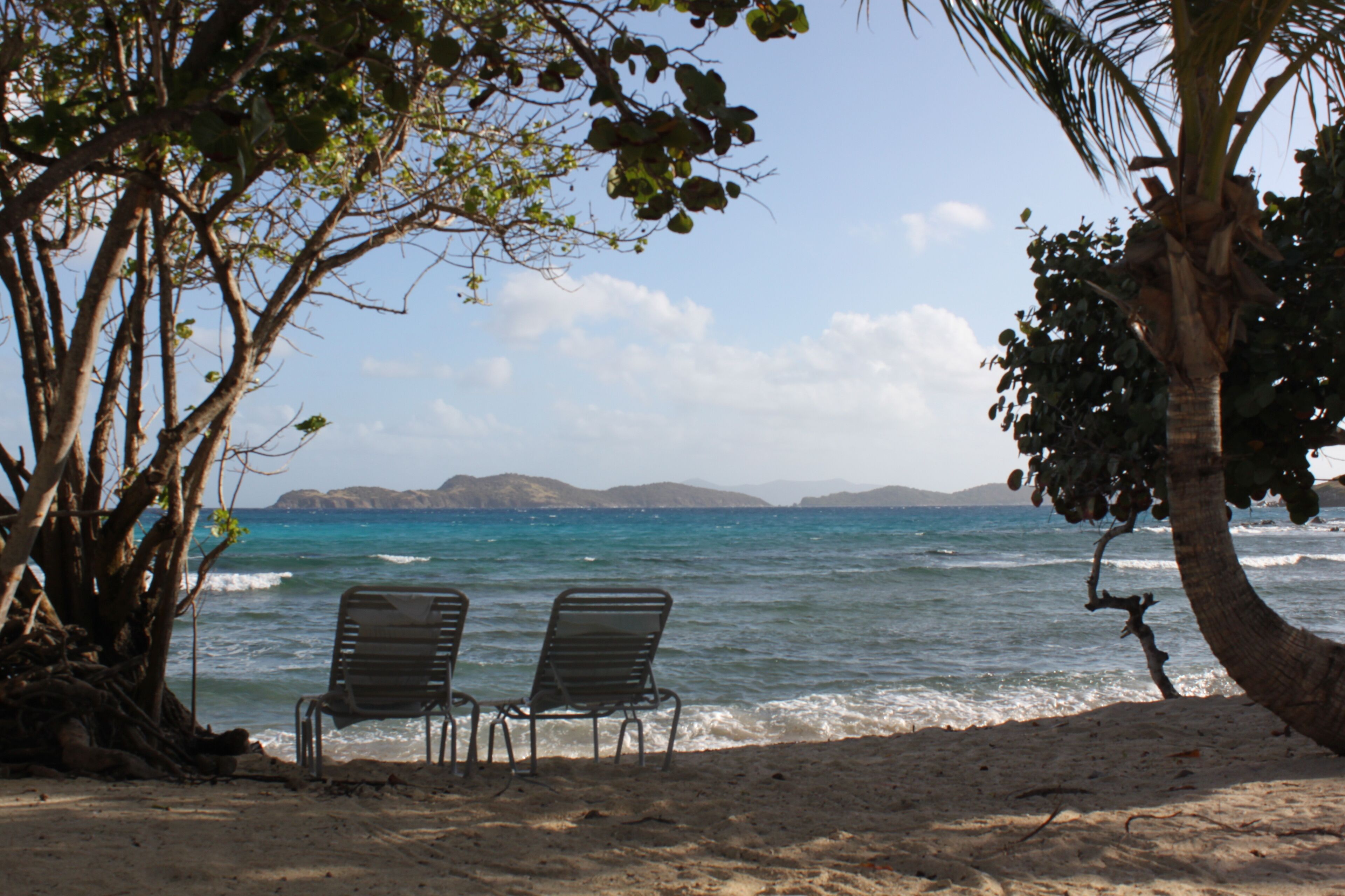 Une plage avec deux chaises en métal, face à l’océan, entourée d’arbres et sous un ciel dégagé.