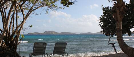 Une plage avec deux chaises en métal, face à l’océan, entourée d’arbres et sous un ciel dégagé.