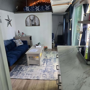 A living room with a blue sofa, a small table, a rug, and a view of the outdoors through a window. - The Little Dipper is back for 2026!  Lake front tiny home w/ private boat dock!  (Andersonville)
