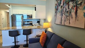 A modern kitchen with white cabinets, a stainless steel refrigerator, and a microwave. There is a bar area with stools and a blue sofa with orange cushions. A painting of palm trees hangs on the wall. - One bedroom one bath oasis! Enjoy one free beach chair set up daily. (Destin)