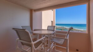 A balcony with a round table and four wicker chairs, overlooking a beach with clear blue water and a sandy shore. - One bedroom one bath oasis! Enjoy one free beach chair set up daily. (Destin)