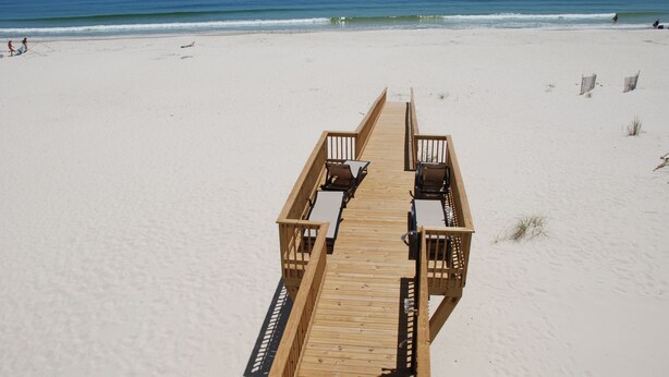 A wooden boardwalk leading to beach chairs and tables on a sandy beach with the ocean in the background. - Luxury Beachfront Home, with Ramp; Sleeps up to 18! (Gulf Shores)