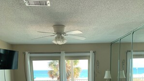 Un salón con un sofá beige grande, una televisión y un ventilador de techo. Hay un ventanal corredero que da a un balcón con vistas al océano y palmeras.