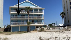 Un hotel frente a la playa con varios balcones, una playa arenosa y un cielo azul despejado.