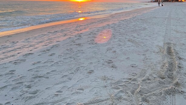 Una playa con huellas, un atardecer y edificios al fondo.