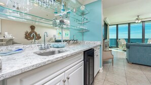 A modern kitchen with a marble countertop, a dishwasher, and a glass shelving unit above the sink. - Phoenix IX 905 - Beautiful 3BR Gulf Front Unit (Orange Beach)