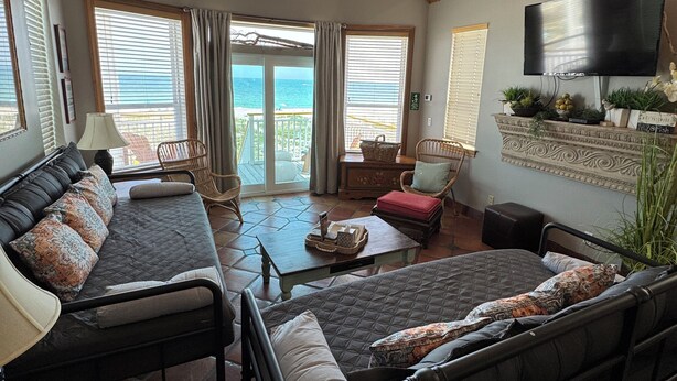 A spacious living room with a high ceiling, a ceiling fan, a large TV, and a view of the ocean through the sliding glass doors. - Beach Front Sleeps 10 Walking distance to everything (St. George Island)