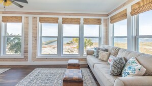 A spacious living room with a large window overlooking a beach, a ceiling fan, and a beige sofa with patterned cushions.