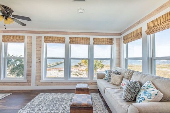 A spacious living room with a large window overlooking a beach, a ceiling fan, and a beige sofa with patterned cushions.