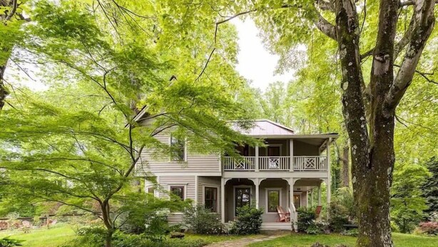 A two-story house with a porch and a covered front entrance, surrounded by lush greenery and trees. - Walk to Main *Outdoor Games* Pet Friendly* Fairy Trail (Saluda)