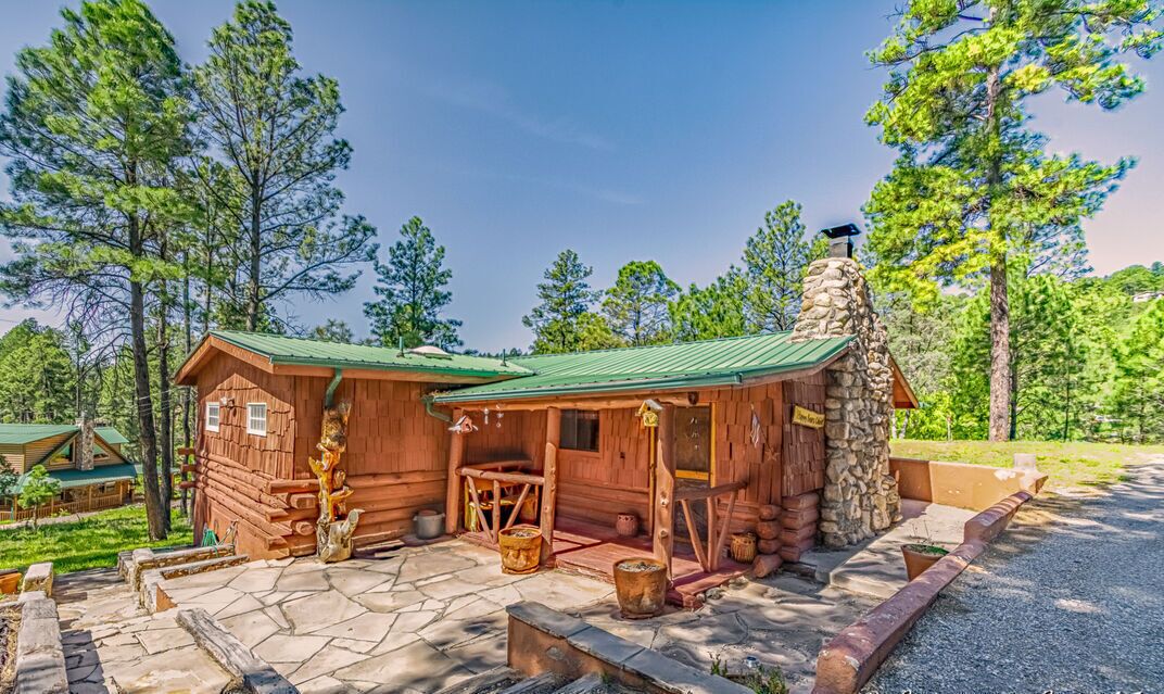 A rustic log cabin with a stone chimney, wooden porch, and a small outdoor seating area.
