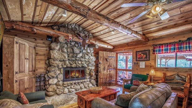 A rustic cabin interior with a stone fireplace, wooden walls, a couch, and a window with a view. - Happy Bears Cabin | Beary Cozy Getaway w/ Fireplace & Forest Views (Ruidoso)