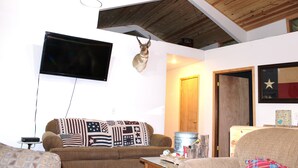 A living room with a mounted television, a sofa with patterned cushions, a wooden ceiling, and a door leading to another room.