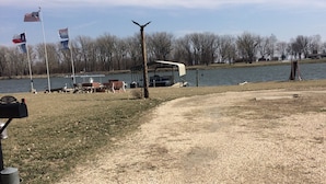 A gravel area with a covered boat ramp, a flagpole with multiple flags, and a body of water in the background.