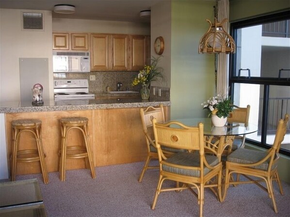 A kitchen with wooden cabinets, a microwave, and a dining area with wicker chairs and a glass table. - SPECTACULAR OCEAN & SUNSET VIEWS!! (Kihei)