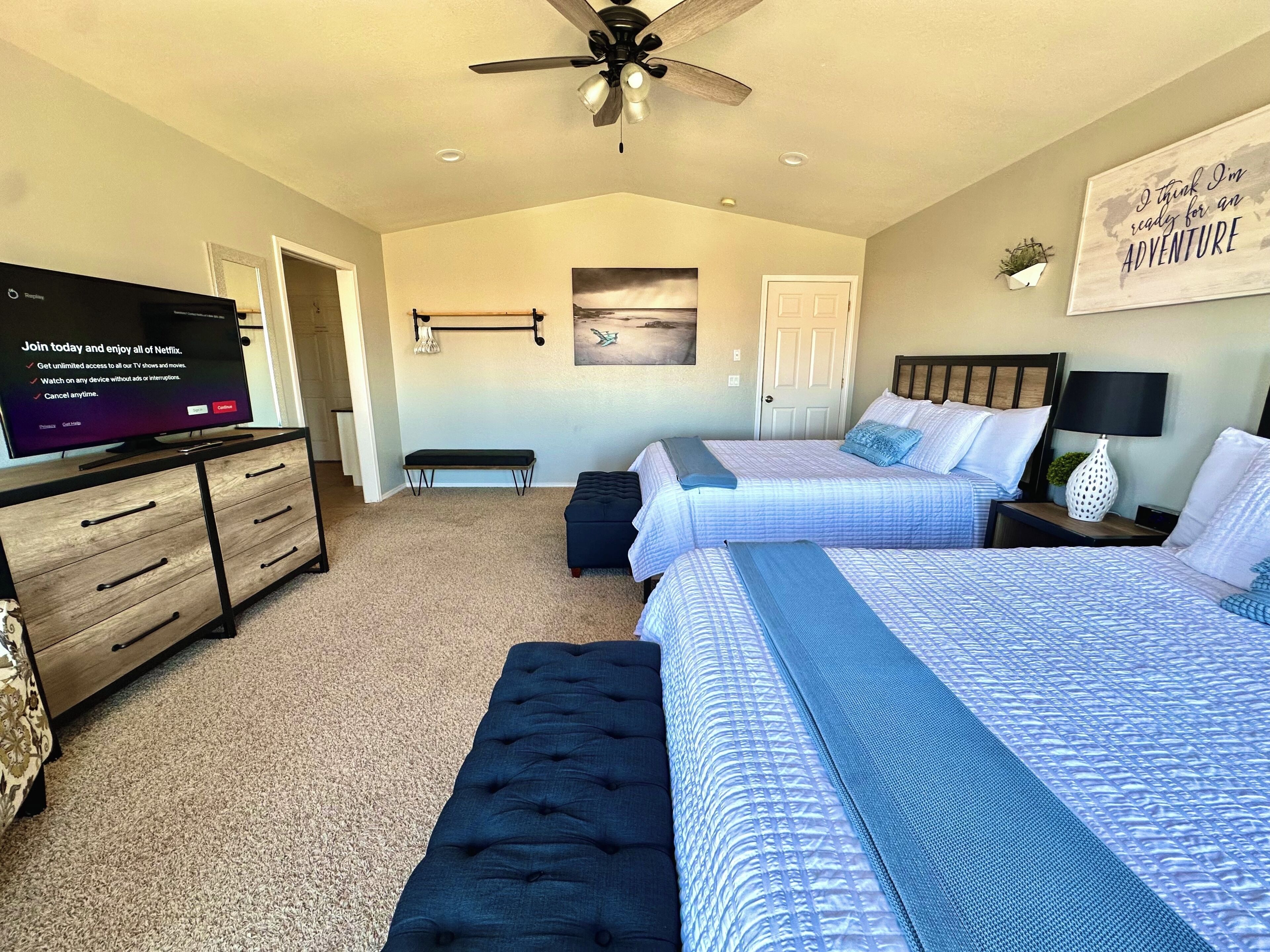 A bedroom with two beds, a TV, a ceiling fan, and a motivational wall art.