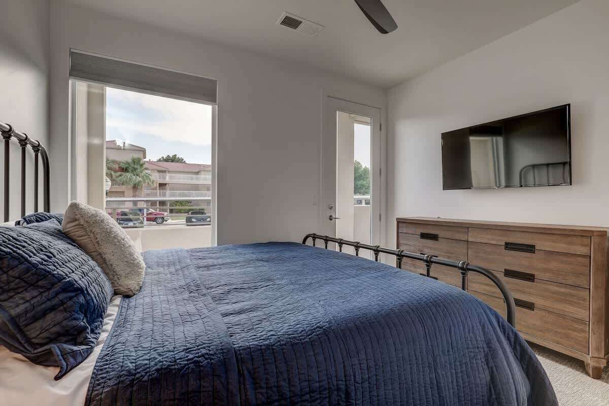 Een slaapkamer met een bed, een aan de muur bevestigde televisie en een raam met uitzicht op gebouwen en geparkeerde auto's.