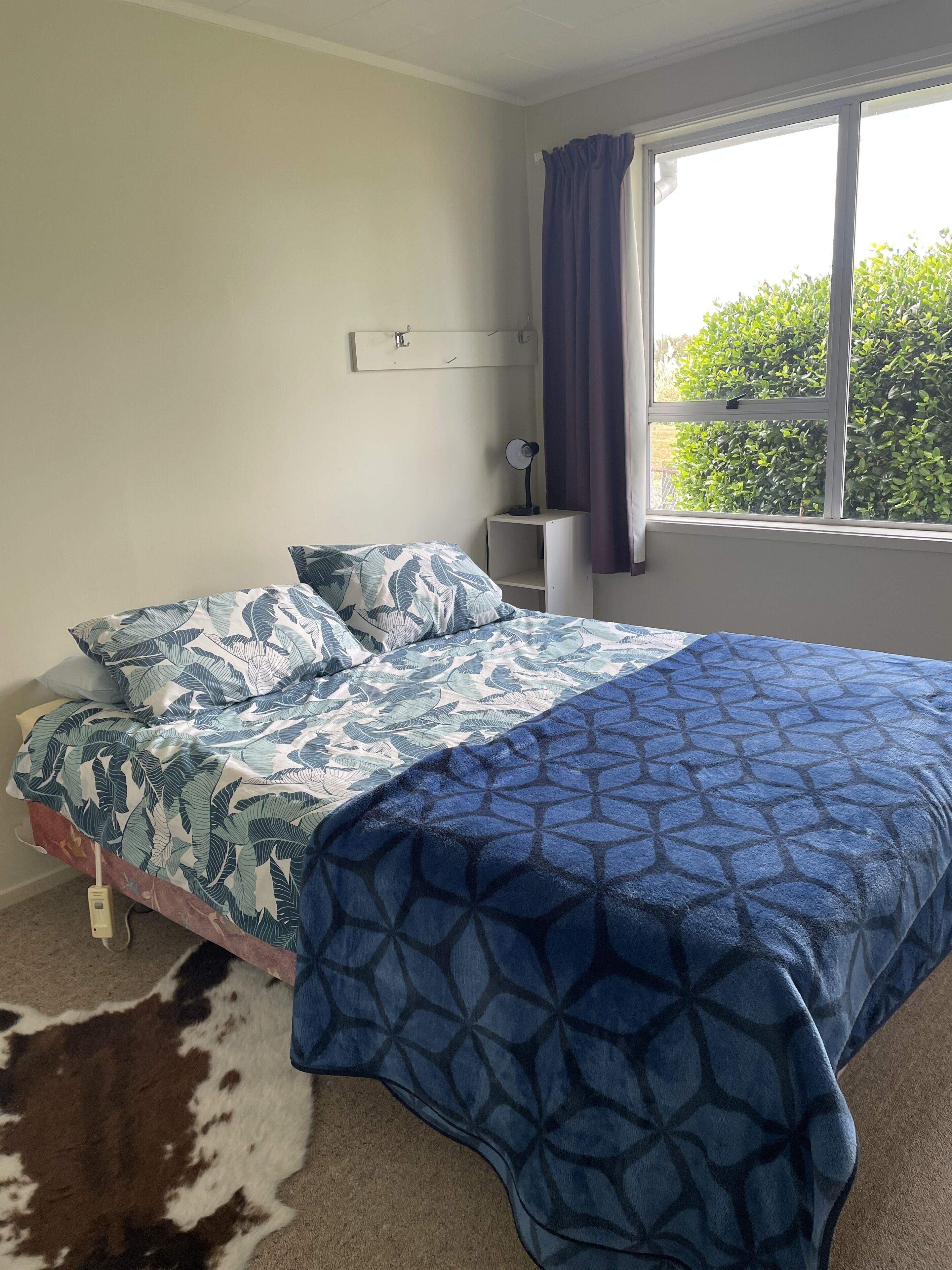 Ein Schlafzimmer mit Bett, Nachttisch, Wandregal, Fenster mit Vorhängen und Blick auf Grünfläche.