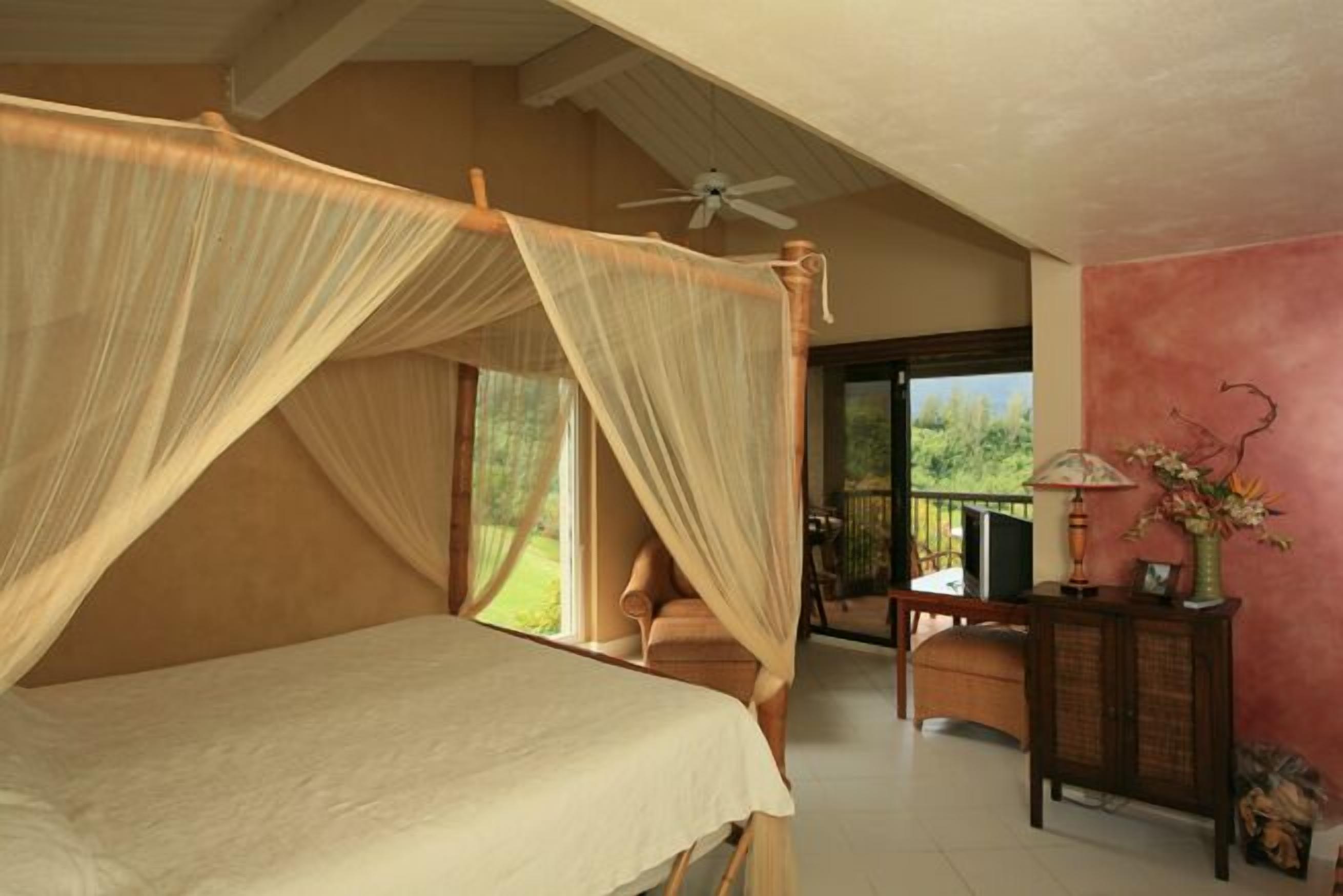 A bedroom with a canopy bed, a ceiling fan, a view of the outdoors, and a small table with a lamp.