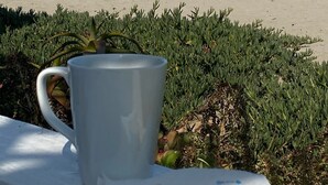 Eine Strandansicht mit einer weißen Tasse, einem Kuchenstück auf einer weißen Decke, einem Rettungsschwimmturm und einem Steg im Hintergrund.