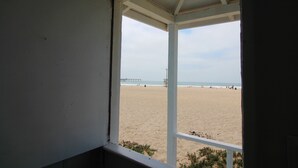 Blick auf einen Strand von einem überdachten Bereich mit weißer Brüstung, ein Steg im Hintergrund und ein Rettungsturm.
