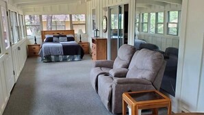 Una habitación alargada y estrecha con una cama, un sofá y una mesita. La habitación tiene ventanas grandes y vistas a árboles.