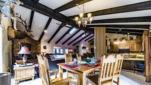 Interior rústico de cabaña con vigas de madera a la vista, una pared de piedra, una zona de comedor con sillas y mesa para cuatro, y una cocina con armarios de madera.