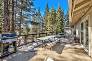 Une terrasse extérieure avec des tables de pique-nique et un barbecue, entourée de grands pins.