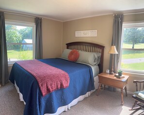 Een slaapkamer met een bed, nachtkastje, lamp, stoel en een raam met gordijnen.