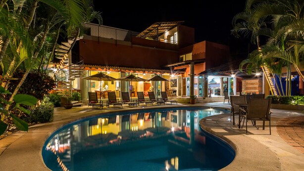 A nighttime swimming pool area with lounge chairs and umbrellas, surrounded by palm trees and a building with a tiled roof.