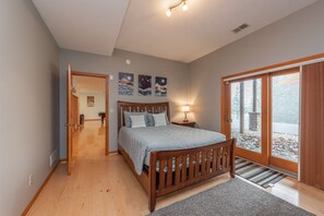 Ein Schlafzimmer mit einem hölzernen Bett, einem Nachttisch mit Lampe, Blick auf einen Wald durch eine Glastür und Kunstwerken an der Wand.