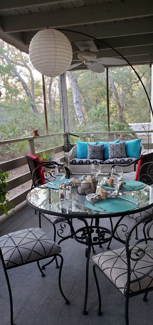 An outdoor patio with a glass table set for dining, surrounded by metal chairs with patterned cushions.