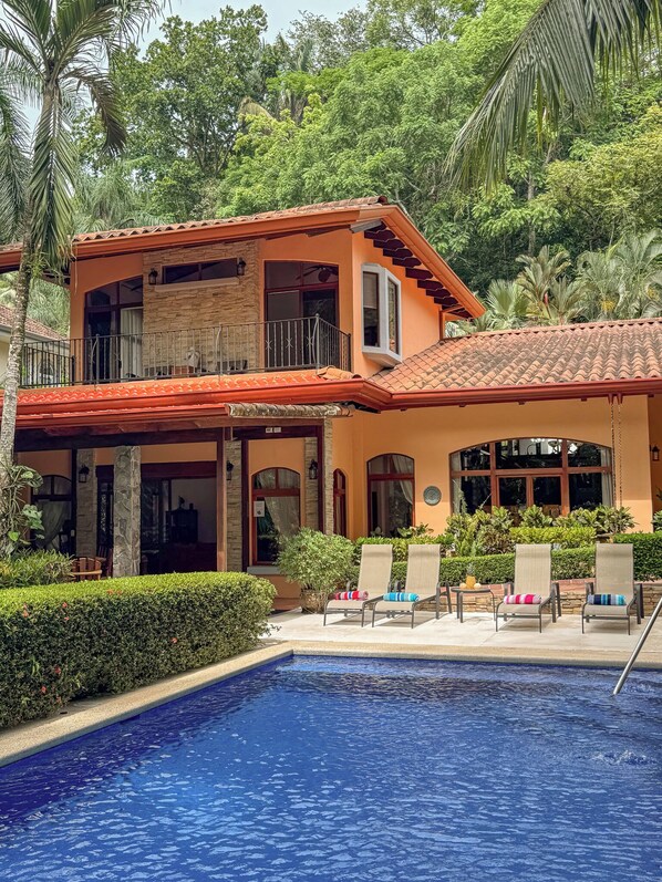 A two-story house with a swimming pool, surrounded by lush greenery.