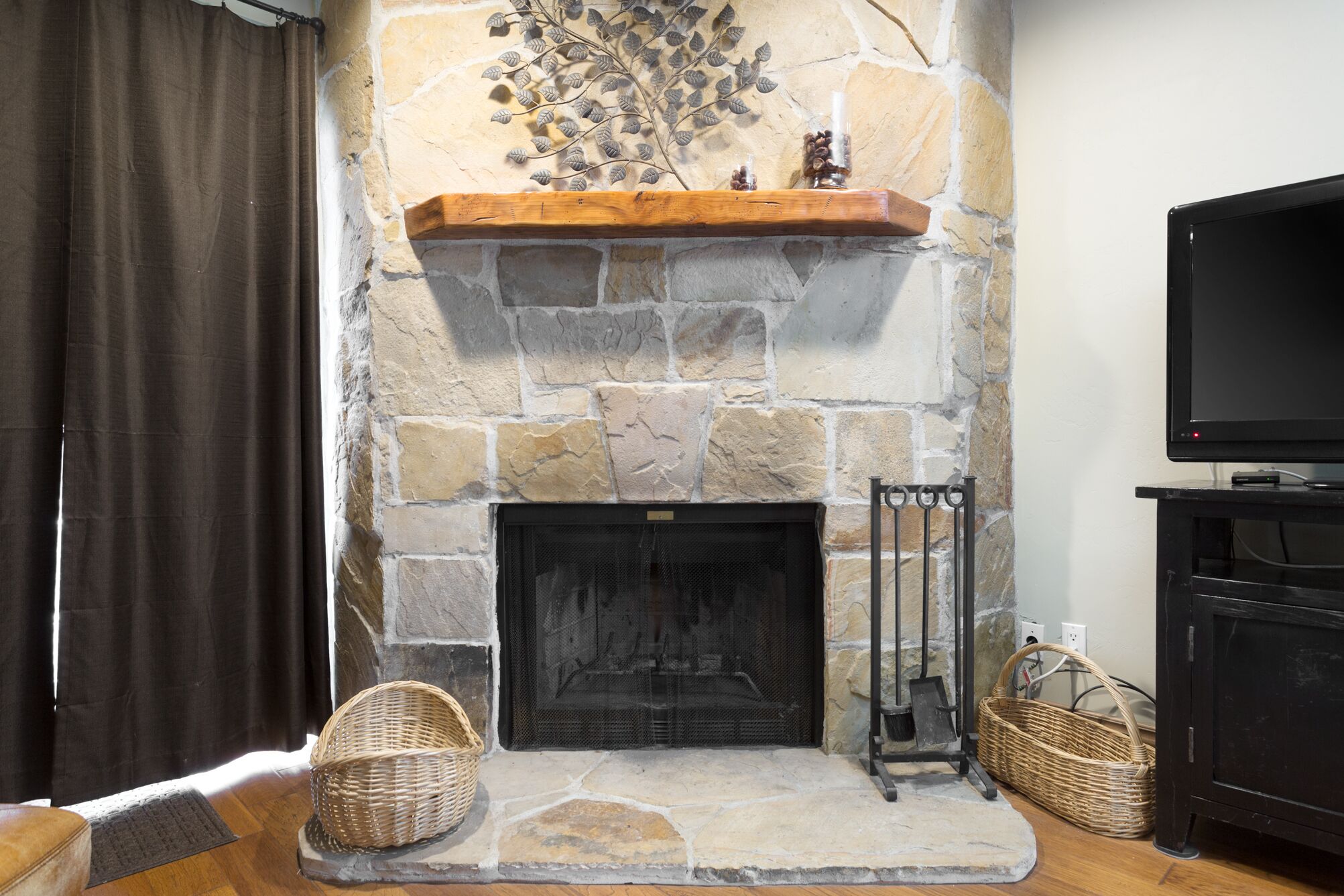 A stone fireplace with a wooden mantel shelf, a television on a stand to the right, and two wicker baskets on the floor.