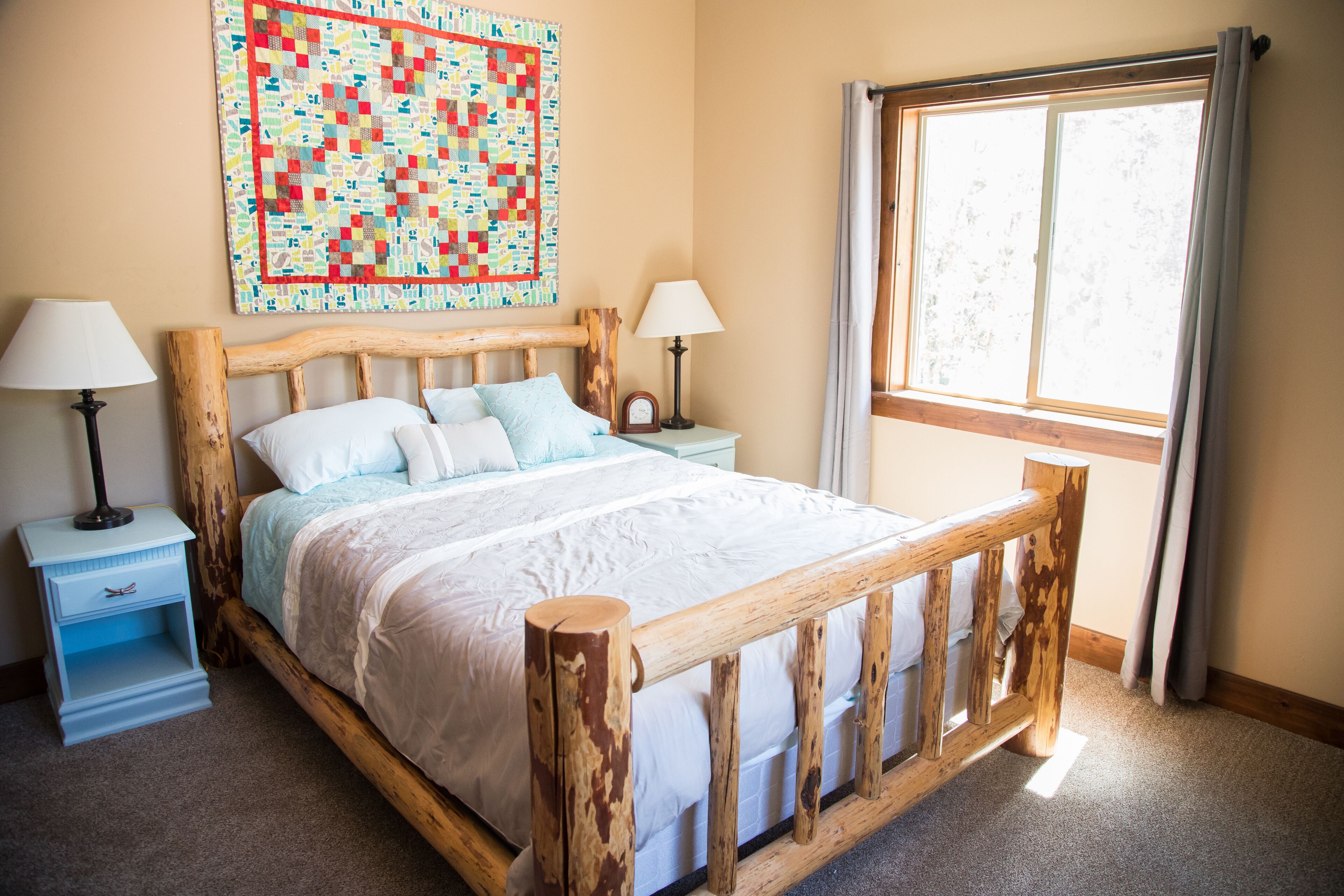 A bedroom with a wooden bed frame, a quilt, two bedside tables with lamps, and a window with curtains.