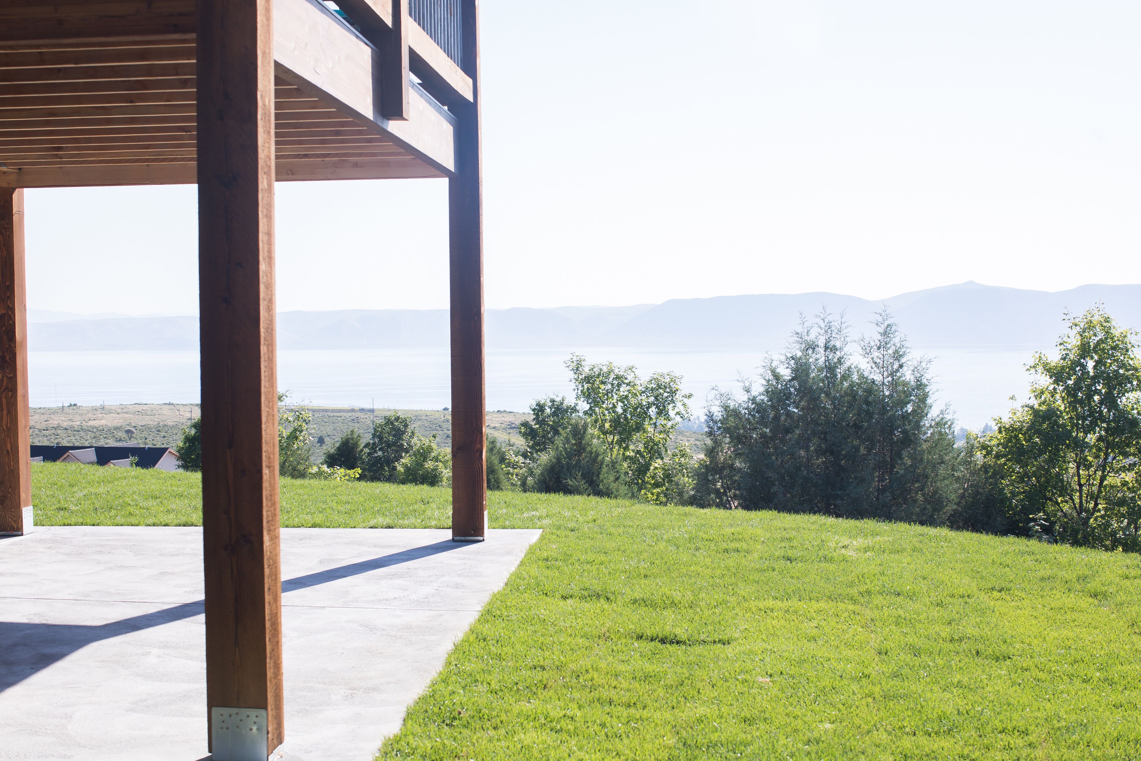 A covered outdoor area with a concrete floor and wooden beams, overlooking a grassy landscape with trees and distant mountains.
