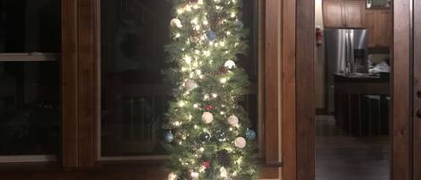 Un árbol de Navidad con luces y adornos, ubicado en una habitación con pisos de madera y ventanas amplias.