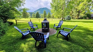 Un espace extérieur circulaire aménagé avec des sièges, doté d’un foyer central et entouré de six fauteuils Adirondack, le tout installé sur une pelouse bien entretenue avec une chaîne de montagnes en arrière-plan.