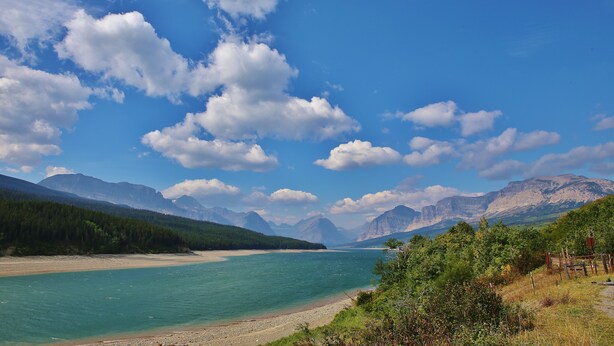Une rivière avec des montagnes en arrière-plan, un ciel dégagé et une zone boisée.