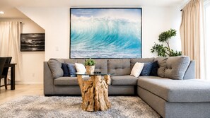 A living room with a grey sectional sofa, a glass coffee table with a wooden base, a large wave painting on the wall, and a plant on the right.
