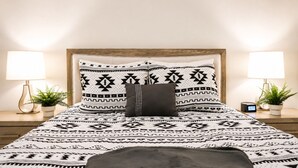 Ein Bett mit gemusterten Betttextilien, zwei Nachttische mit Lampen, eine Uhr und eine Pflanze auf jeder Seite.