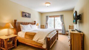 Een houten bed met witte lakens, een nachtkastje met een lamp, een televisie op een houten dressoir en een raam met gordijnen.