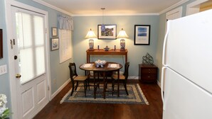 A dining area with a wooden table, chairs, and a rug. There are two wall-mounted lamps, a framed picture, and a refrigerator. - QUAINT COTTAGE IN HISTORIC OLD POINT   Beaufort, S.C. (Beaufort)