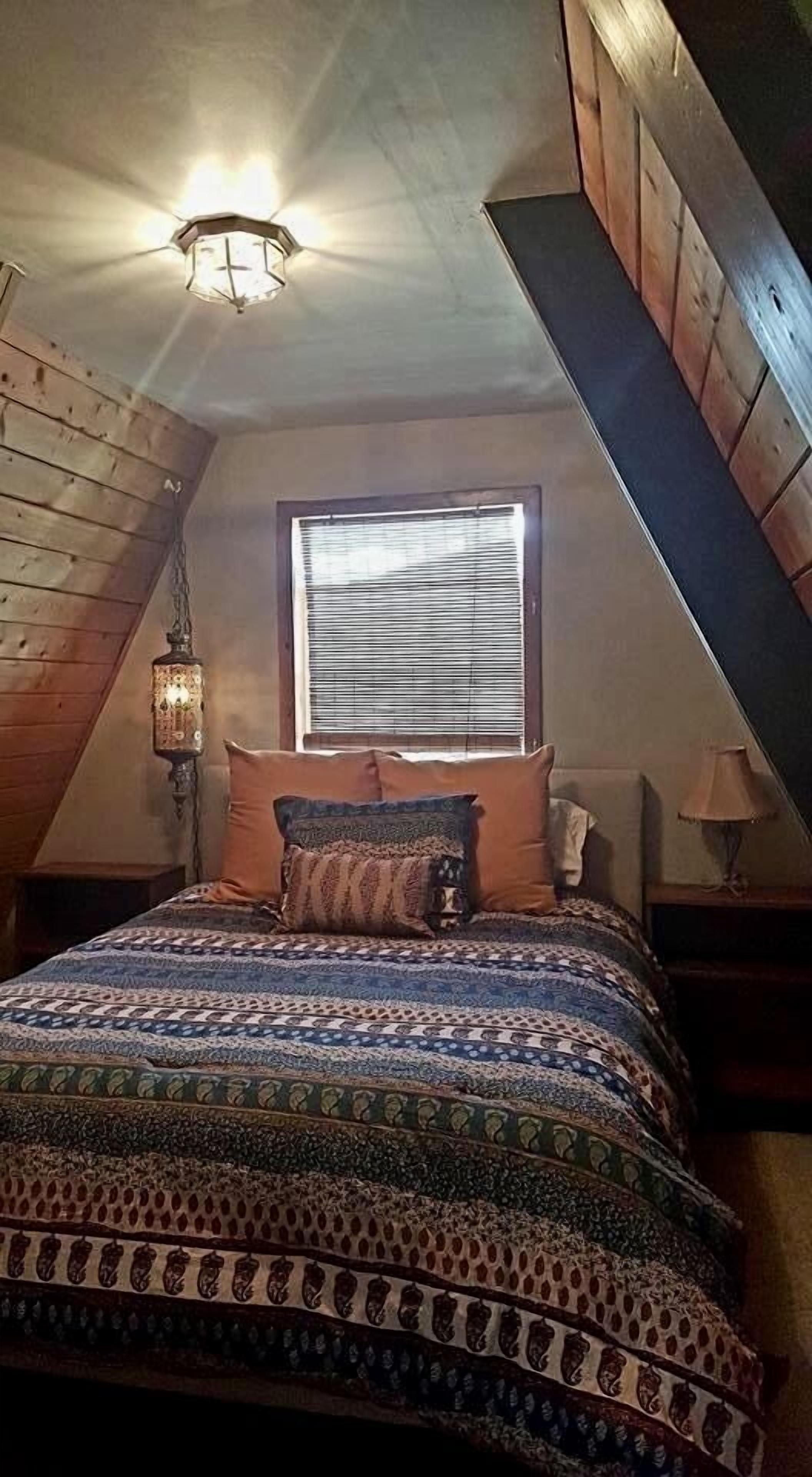 Un dormitorio con techo inclinado, una ventana con persianas, una colcha estampada y un farol colgante.